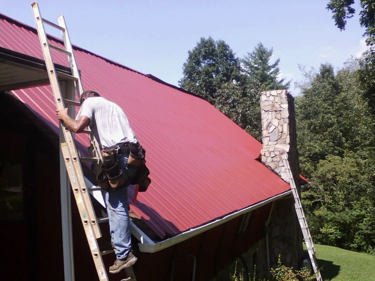Roofer on a ladder beside a red metal roof and stone chimney — Soffit & Fascia Repair by Roofing Brothers in Falls City